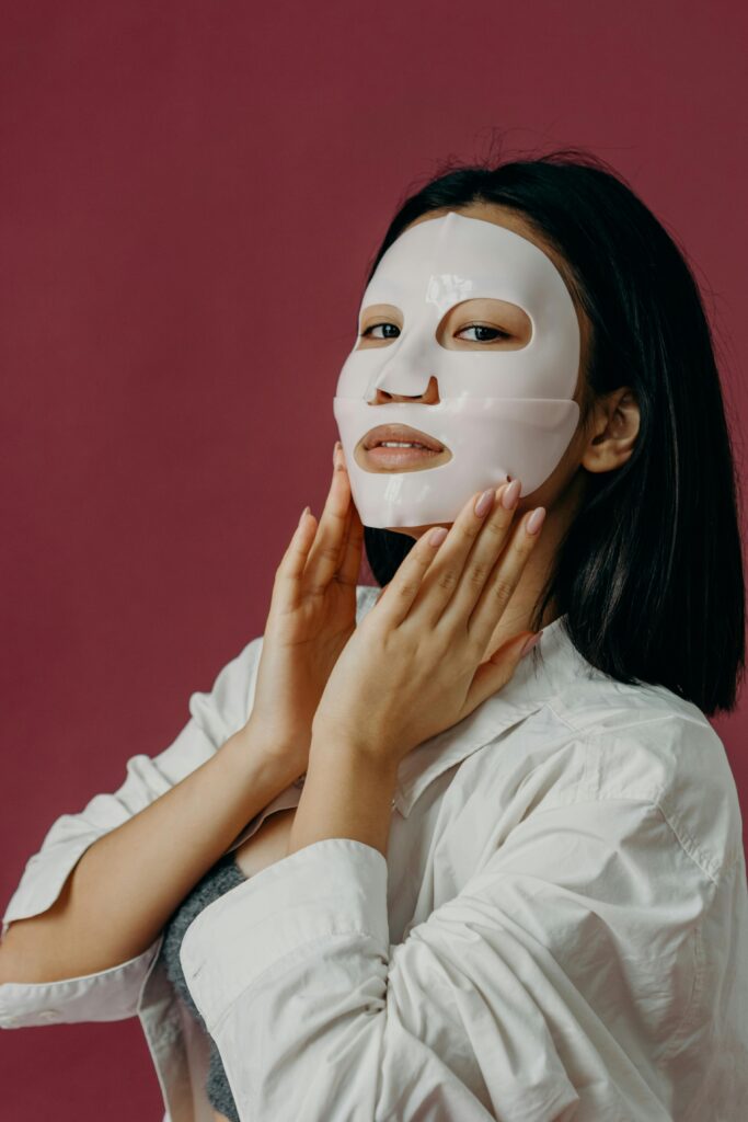 Asian woman wearing a sheet mask for skincare routine in studio setting with red background.